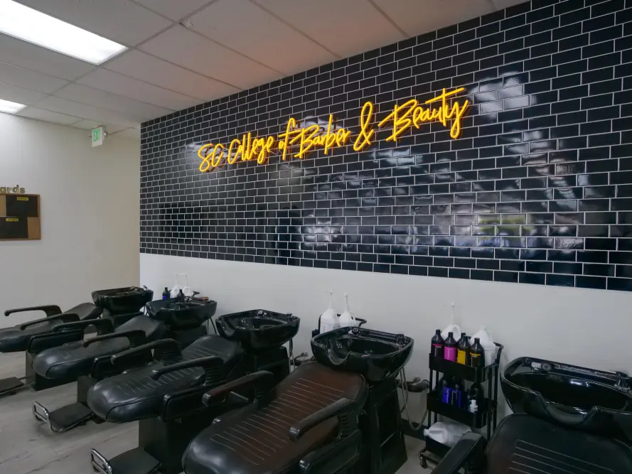 Interior of the Southern California College of Barber & Beauty salon with rows of black shampoo chairs, wash basins, and hair products beneath a black brick wall bearing the school’s yellow script sign.