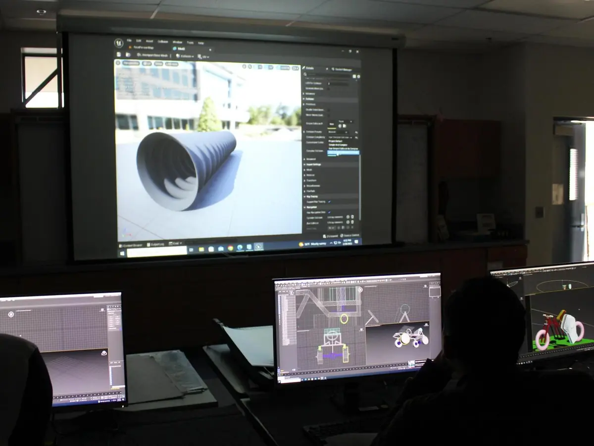 Students work at desktop computers in a dark classroom while a large projection screen displays 3D modeling software.