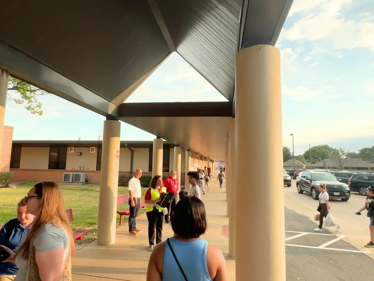People walk beneath the covered entrance walkway of a low brick school building beside a parking lot.