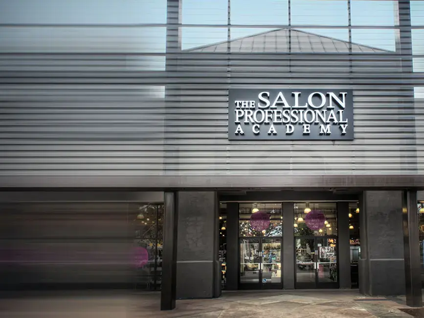 Exterior entrance of The Salon Professional Academy, with the academy name displayed above the glass doors on a dark horizontal-paneled facade.