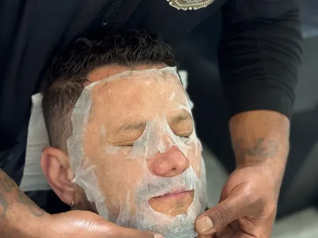 A cosmetology student applies a facial treatment to a seated man while wearing a Universal College of Beauty uniform.