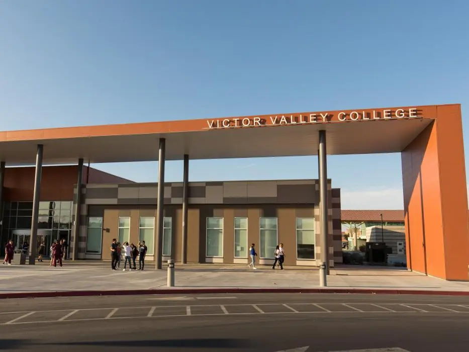 Front entrance of a Victor Valley College building, with the college name displayed across the orange roofline and several people standing near the entrance.