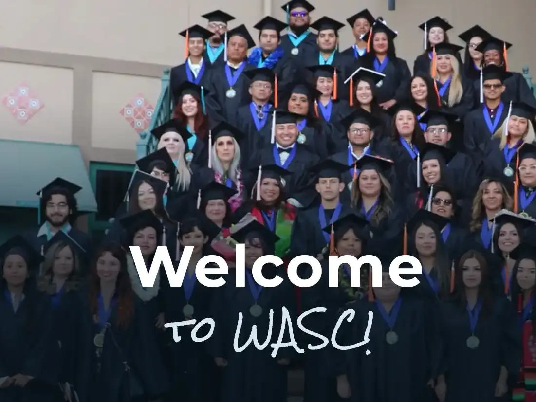 Large group of graduates wearing black caps and gowns gathered on an indoor staircase beneath text reading “Welcome to WASC!”