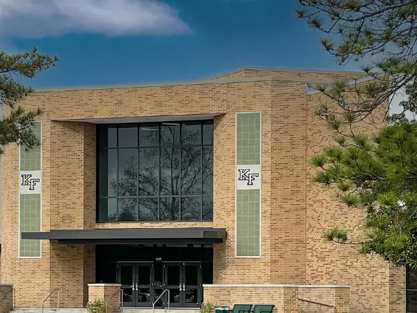 Front entrance of Klein Forest High School, a tan brick building with a tall central glass facade, wide entry doors, trees, a driveway, and a grassy lawn.