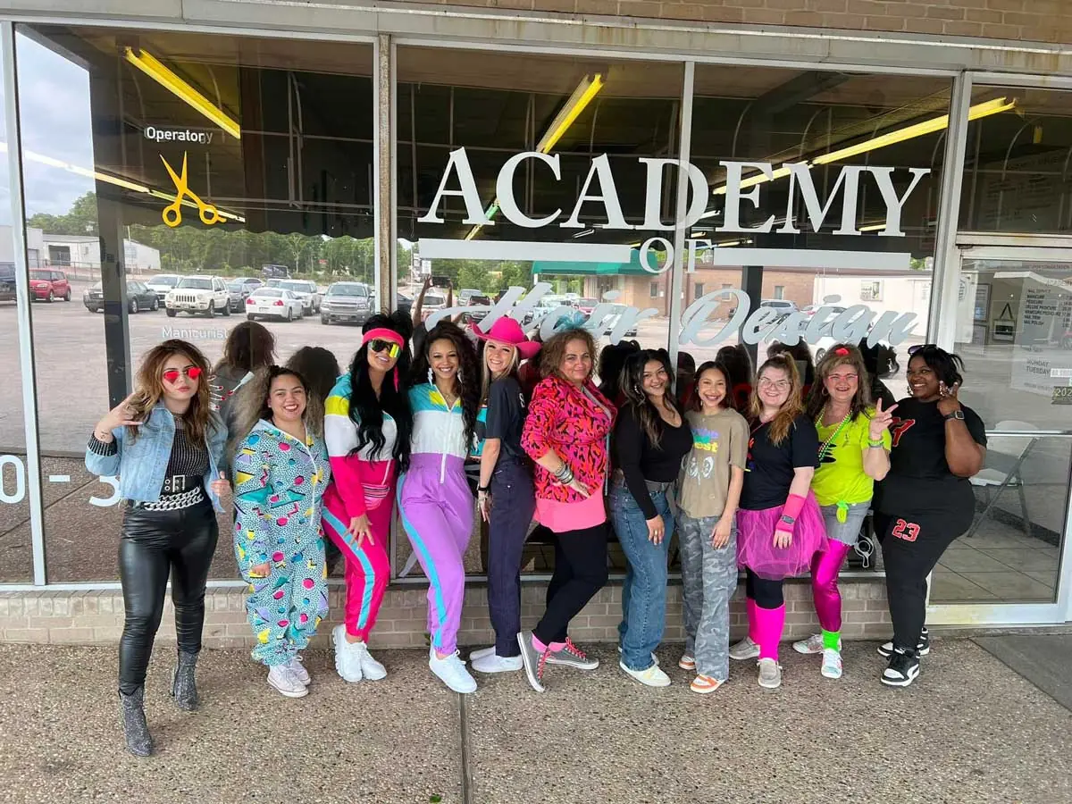 A group of people stands outside the glass storefront of the Academy of Hair Design, with large white ACADEMY lettering and hair-design graphics on the windows.