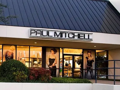 Exterior of a Paul Mitchell salon school in a shopping-center building, with a large PAUL MITCHELL sign above glass windows and doors, landscaped shrubs, and hair-model posters in the windows.