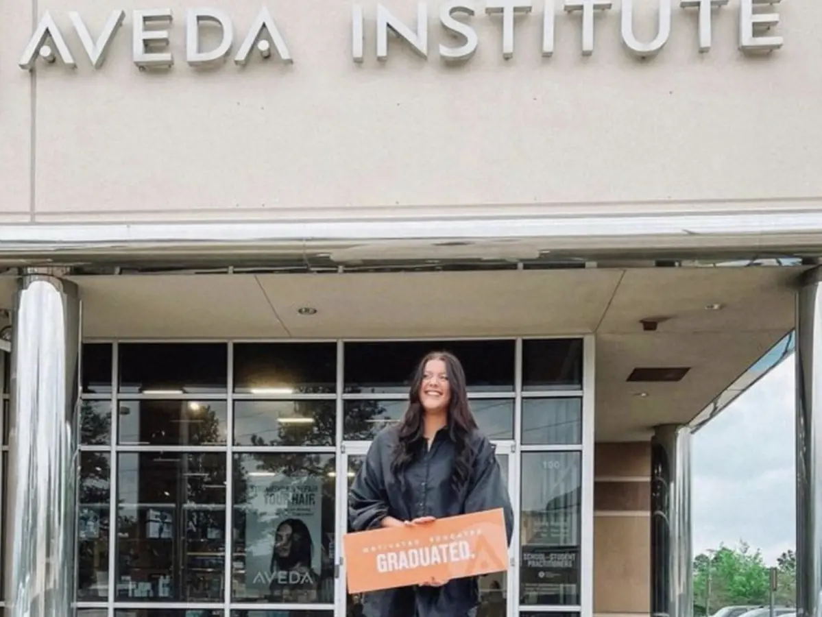 A person stands outside the glass entrance of the Aveda Institute, holding a graduation sign beneath the building's Aveda Institute lettering.