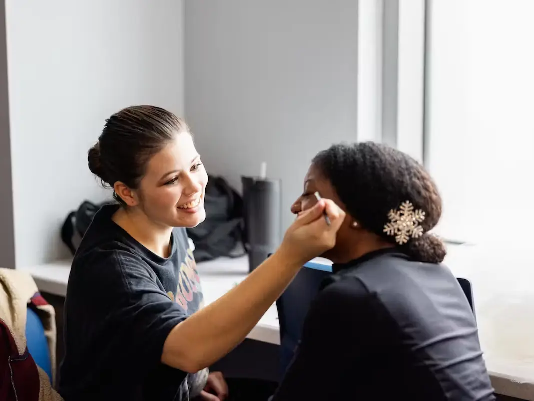 A cosmetology student applies makeup to a seated client in a bright salon classroom, with styling equipment visible behind them.