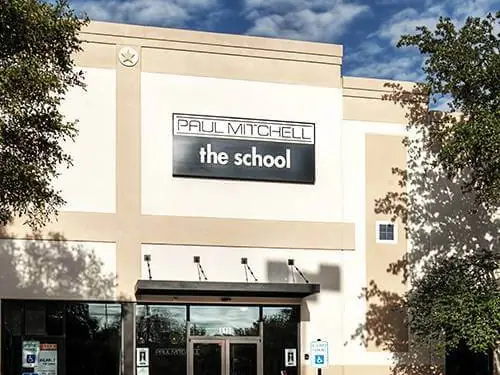 Front exterior of Paul Mitchell the school in a beige commercial building, with the school sign above glass entrance doors, trees on both sides, and a cloudy blue sky.