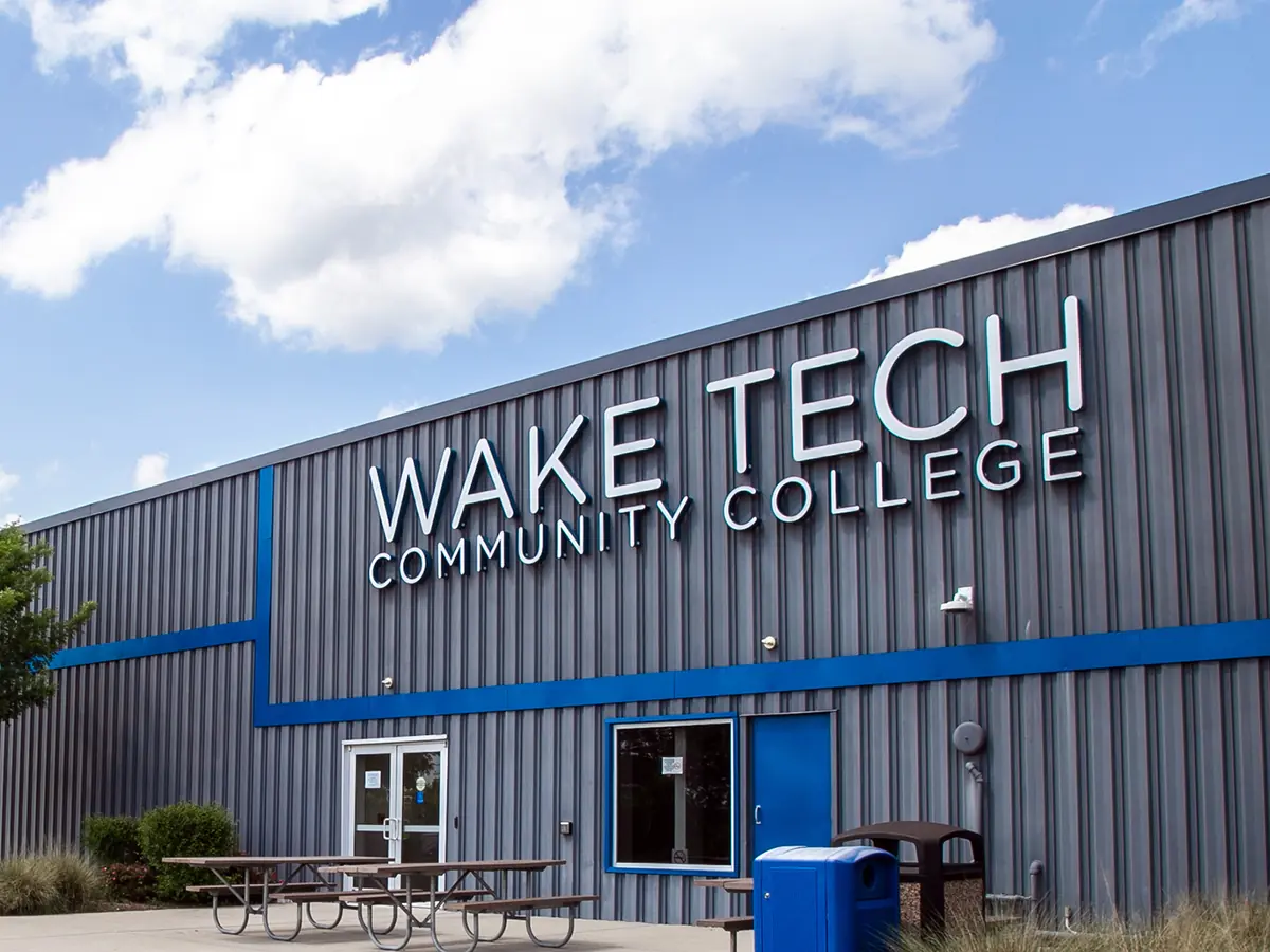 Exterior of Wake Tech Community College's Beltline Education Center, a dark gray building with blue accents, large Wake Tech signage, entrance doors, trees, and picnic tables under a partly cloudy sky.