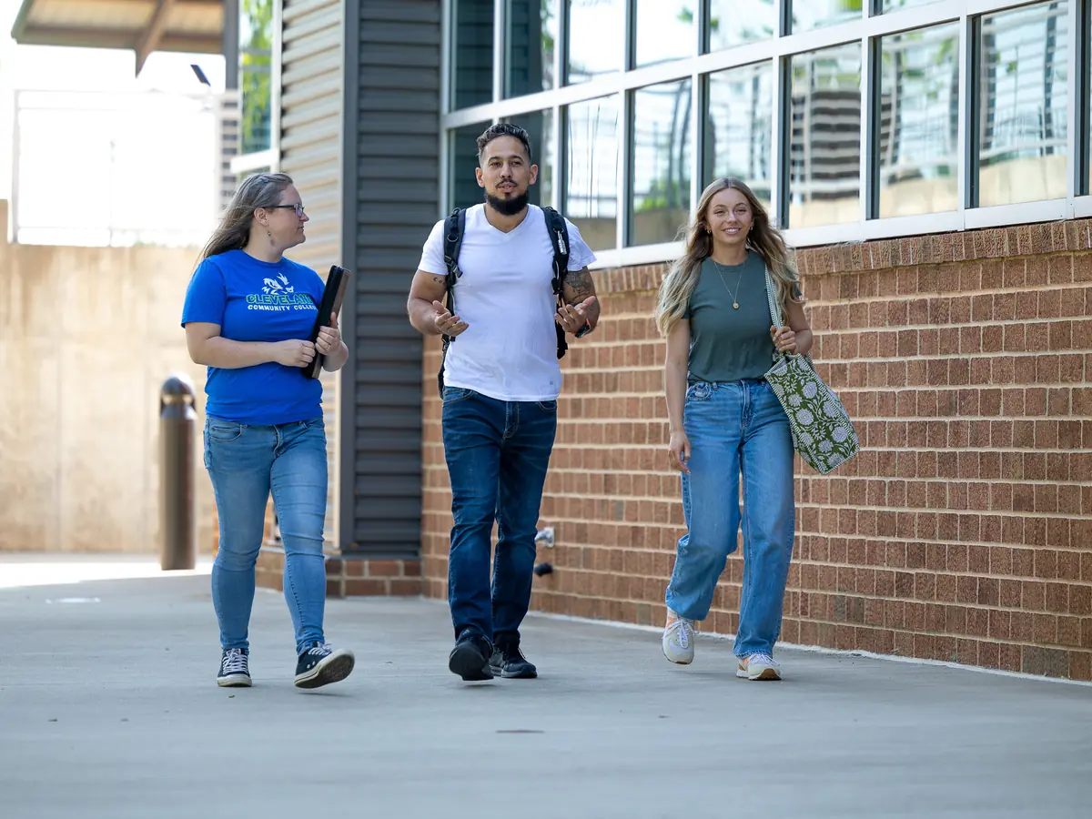 Three students walk along a covered campus walkway beside a brick-and-glass building.