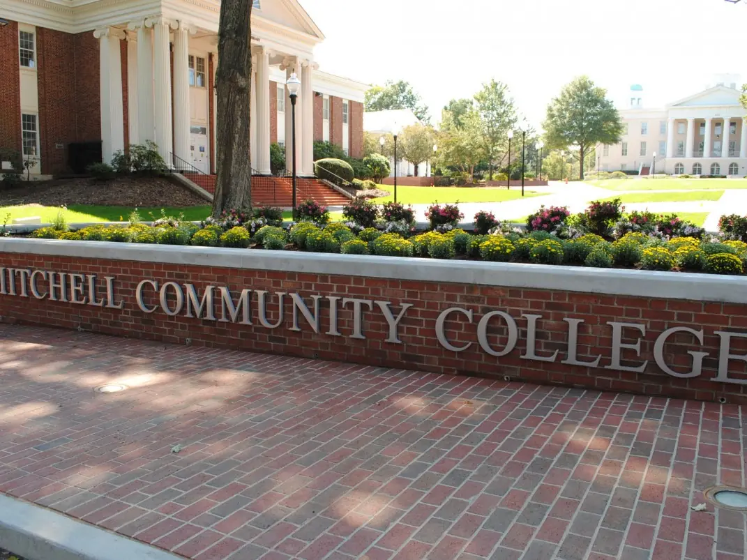 Brick campus entrance sign reading “MITCHELL COMMUNITY COLLEGE” in front of landscaped college grounds and campus buildings.