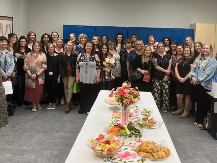 A large group of people poses around a flower-decorated refreshments table inside the WCC Ashe Campus School of Cosmetic Arts, with framed hair-themed artwork on the walls.
