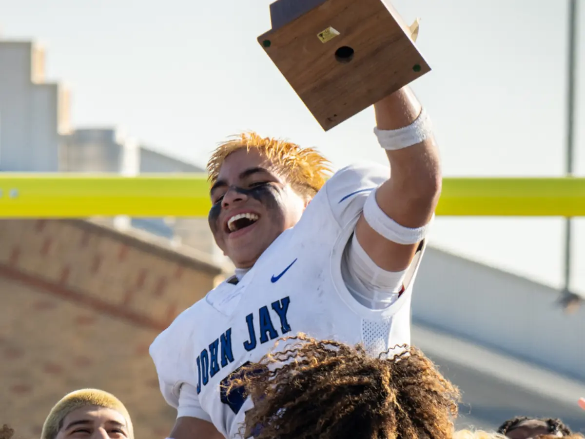 A student in a white John Jay T-shirt raises a graduation cap above a crowd outdoors, with a yellow goalpost and school buildings behind them.
