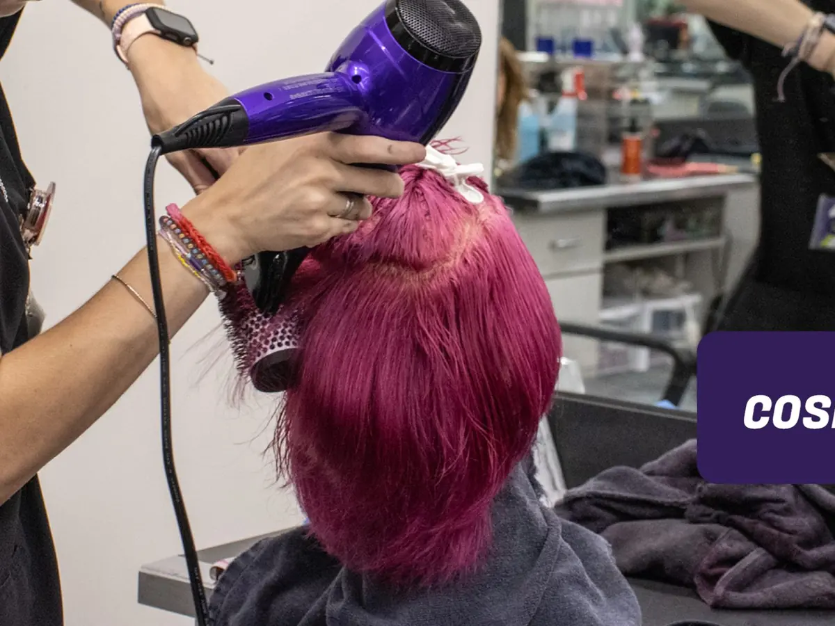 A cosmetology student uses a purple hair dryer and brush to style a bright pink mannequin head in a salon classroom, with another mannequin and salon stations in the background.
