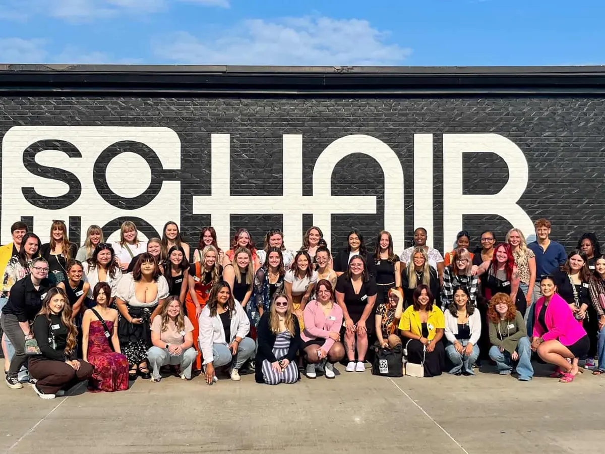 A large group of people poses outside in front of a black brick wall painted with the word “SOHAIR.”