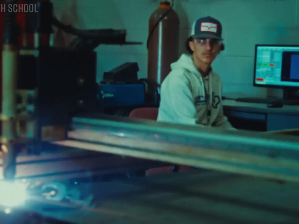 Student wearing a cap seated in a technical workspace beside computer equipment and illuminated machinery.