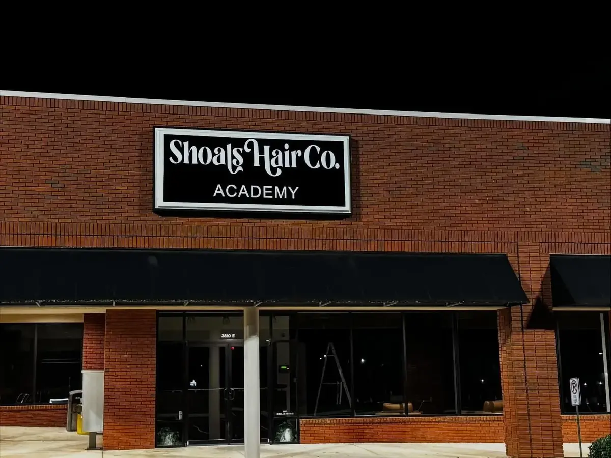 Exterior of a brick building with a black sign reading “Shoals Hair Co. Academy” above the storefront entrance.