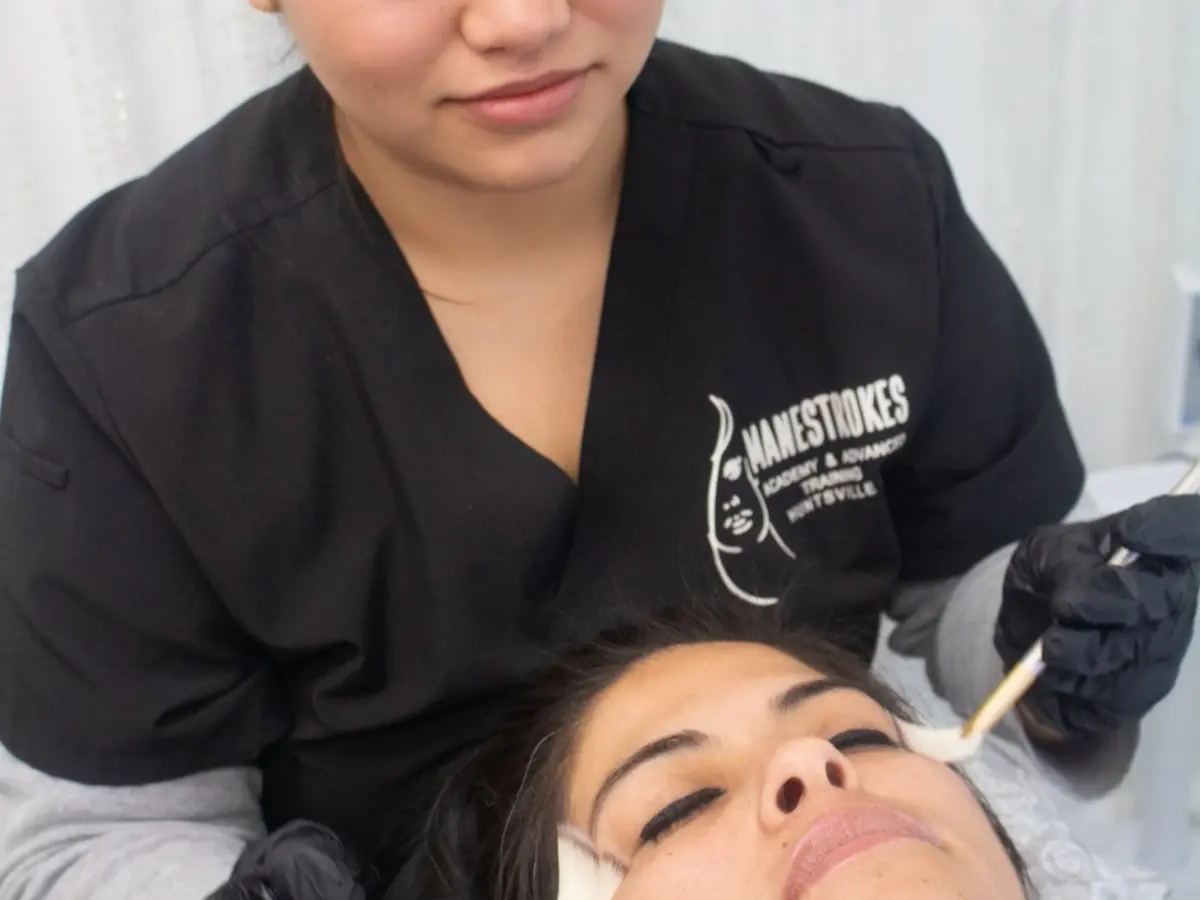 A cosmetology student in a black Mane Strokes Academy uniform applies a treatment to a client's face while the client reclines in a salon chair.