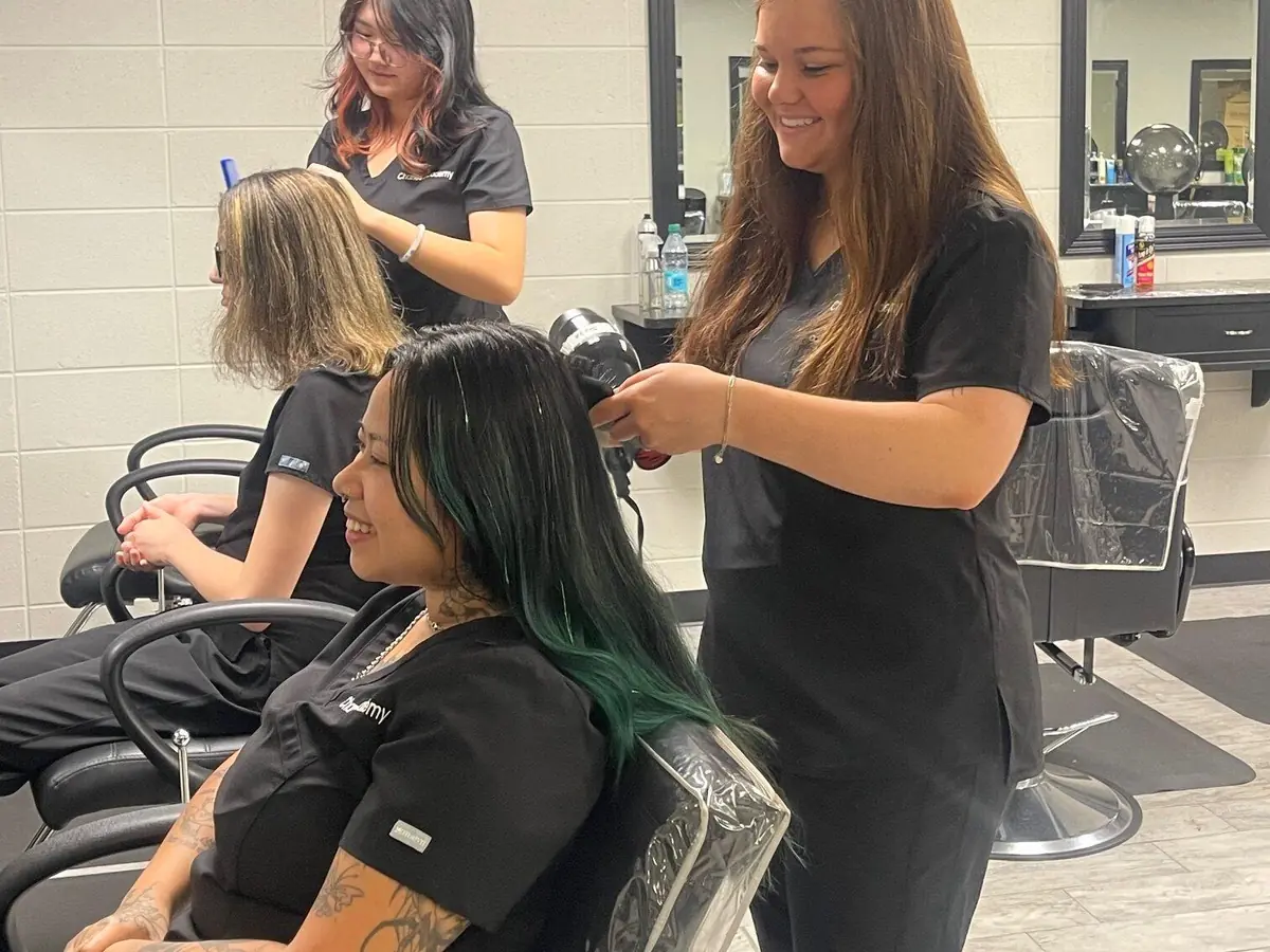 Two hairstylists work on clients seated in salon chairs inside a cosmetology salon, with mirrors and styling stations along the wall.