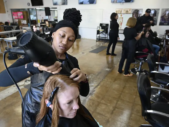 Students practice hairstyling in a cosmetology school salon, with a student blow-drying a seated client in the foreground and additional styling stations behind them.