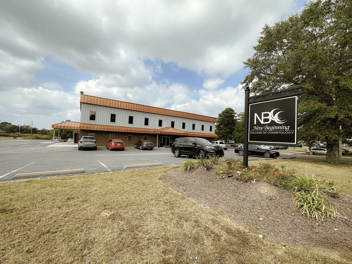 Exterior of New Beginning College of Cosmetology, showing the long campus building, parking lot, and roadside NBCC sign.
