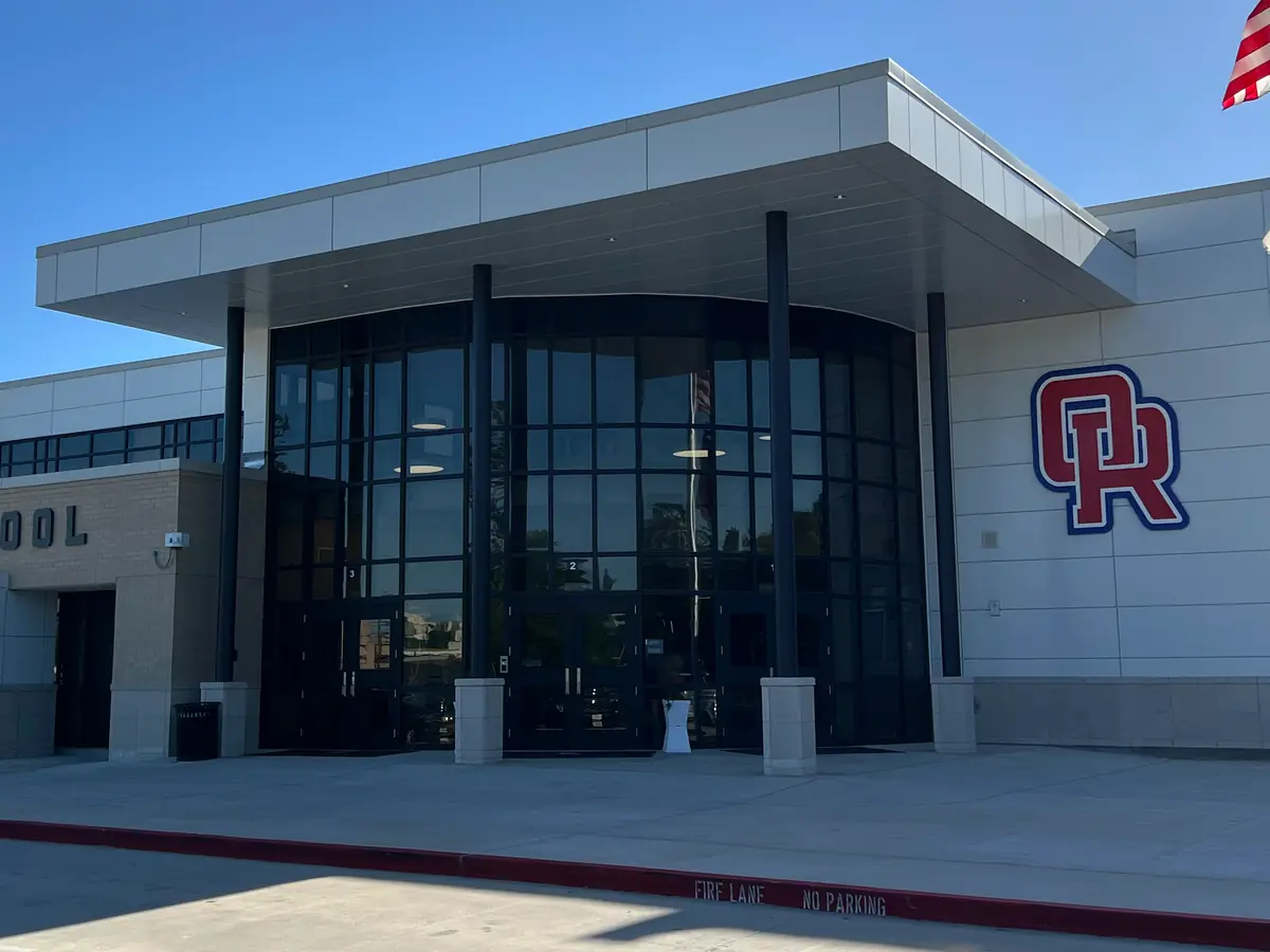 Front exterior of Oak Ridge High School, showing the glass entrance, Oak Ridge High School lettering, OR logo, and Texas and U.S. flags under a blue sky.