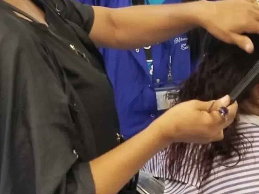 A barbering student in a striped shirt receives a haircut from an instructor wearing a black salon cape in an indoor training area.