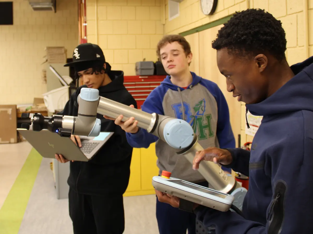 Three students work together with a small industrial robotic arm and metal components in a vocational technology classroom.
