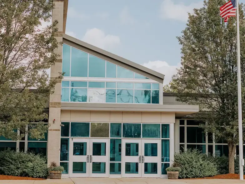 Front entrance of Greater Lawrence Technical School, showing the glass-fronted entry beneath a sloped roof, trees, flagpole, and doors.