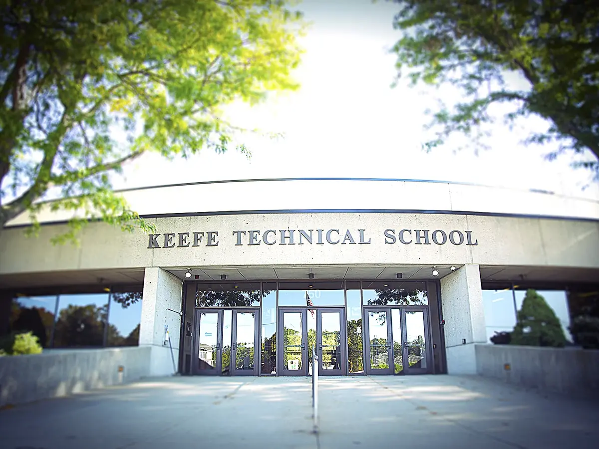 Front entrance of Keefe Technical School with glass doors beneath a facade labeled “KEEFE TECHNICAL SCHOOL,” framed by trees.