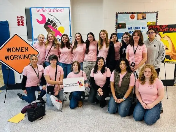 A group of students and adults in pink shirts pose indoors beside a sign reading Lower Pioneer Valley Career and Technical Center, with a Women Working road sign and event displays behind them.