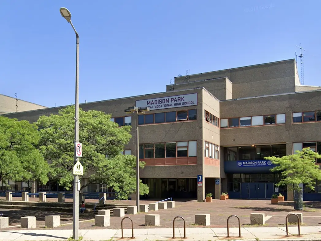 Exterior of Madison Park Technical Vocational High School, a broad concrete school building behind trees and a paved forecourt with bicycle racks.