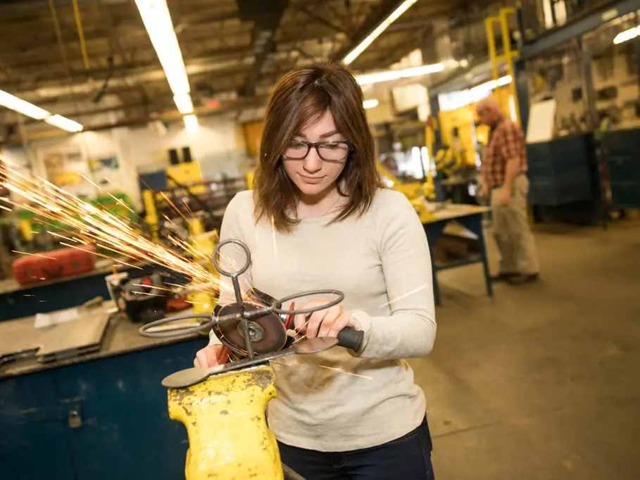 Student wearing safety glasses uses a handheld grinder on a metal object in a vocational metalworking shop, with sparks flying and industrial equipment in the background.