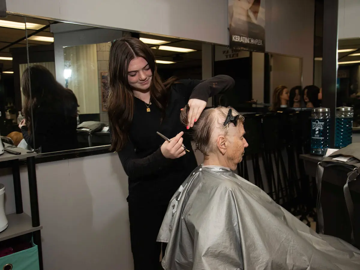 A student in a cosmetology classroom trims a seated client's hair beside mirrored styling stations and salon equipment.