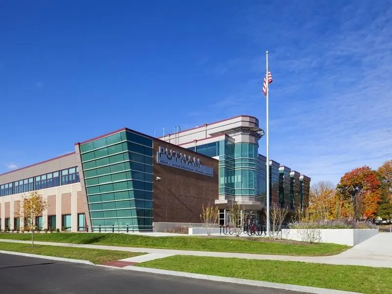 Exterior of Roger Putnam Vocational Technical High School, a modern brick-and-glass building with a flagpole, broad sidewalk, lawn, and autumn trees.