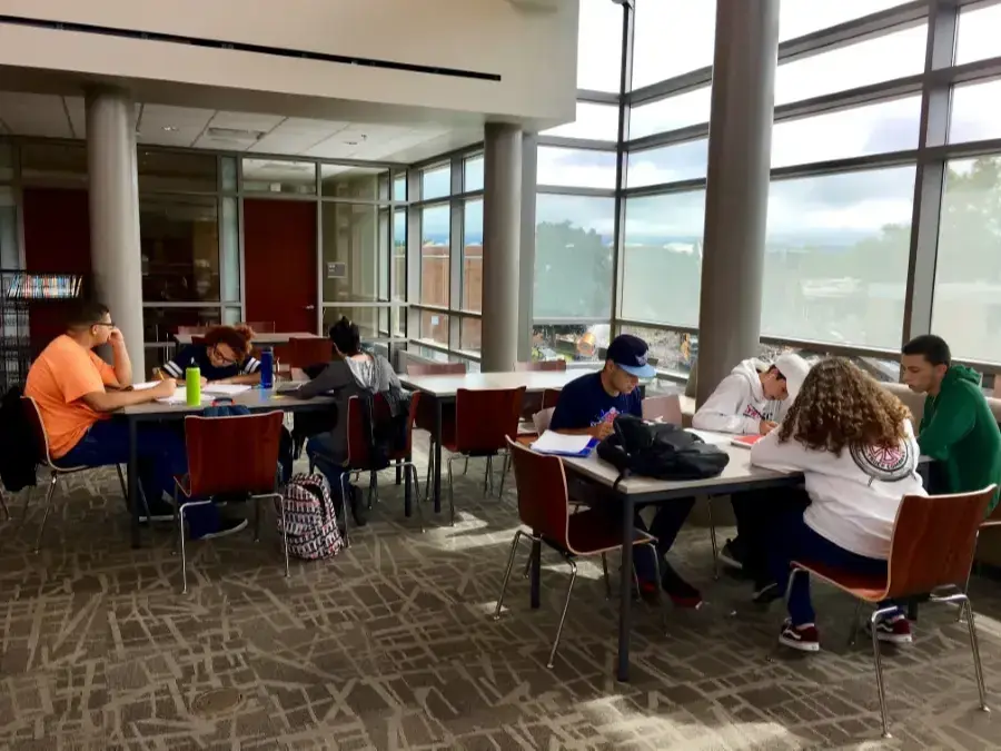Students sit at tables and study in the bright Taconic High School library, which has floor-to-ceiling windows, white columns, and red chairs.