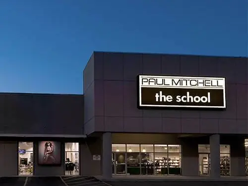 Exterior of Paul Mitchell the school in a shopping-center storefront at dusk, with the illuminated school sign above the entrance and salon windows below.