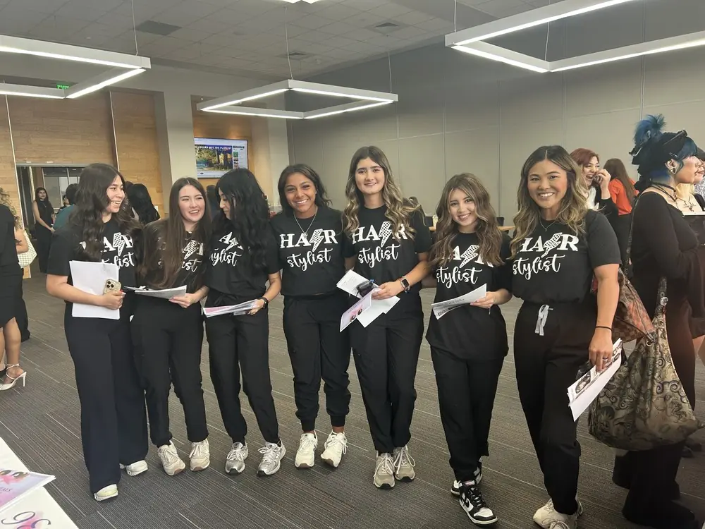 A group of cosmetology students in black shirts stand together in a brightly lit salon-style room, with mirrors and other people visible behind them.
