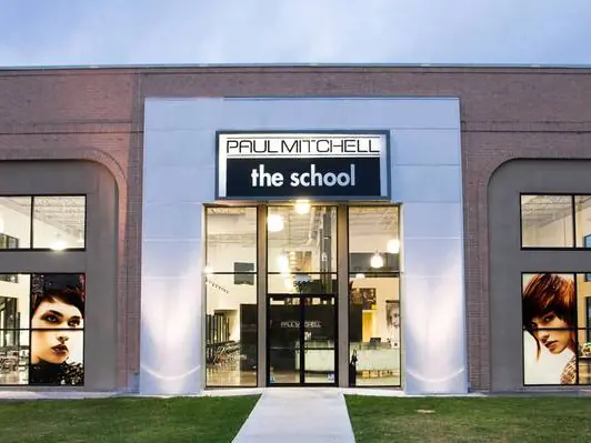 Front exterior of a single-story Paul Mitchell The School building at dusk, with large windows, a centered glass entrance, and the school's name displayed above the doors.