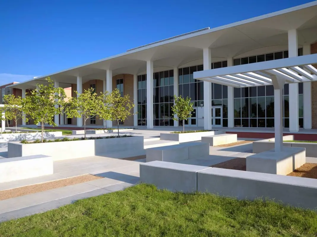 Exterior of a modern school building with tall white columns, large glass windows, a covered entrance canopy, concrete seating walls, young trees, and landscaped grass under a clear blue sky.