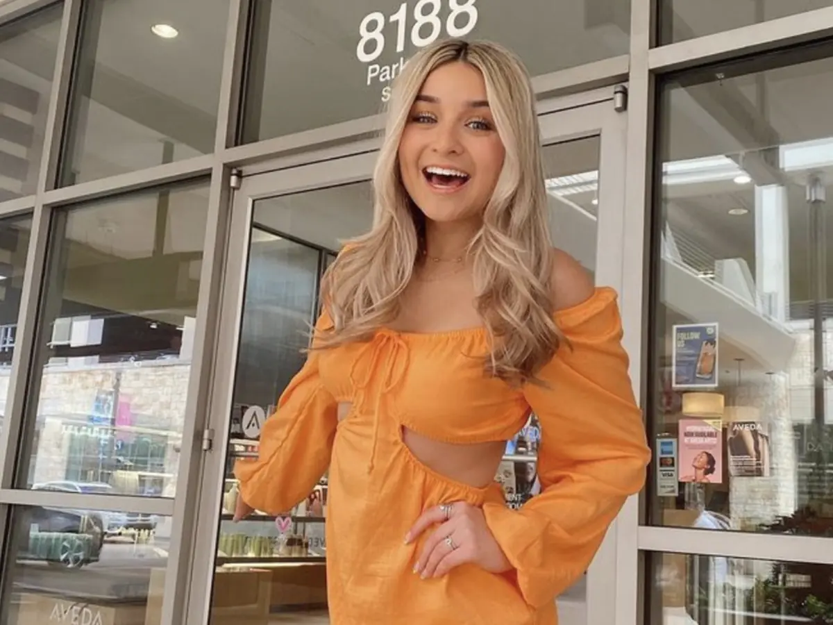 A smiling woman in an orange outfit stands in front of the Aveda entrance, where “8188” and partial “Park…” lettering are visible on the glass doors.