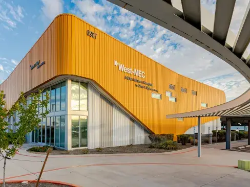 Exterior of a West-MEC campus building with a curved orange facade, large West-MEC signage, glass entrance, covered walkway, and desert landscaping.