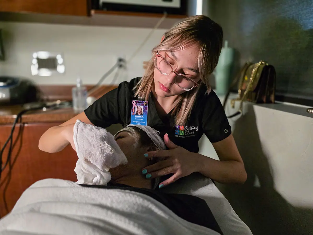 Cosmetology student wearing a black Cutting Class uniform performs a treatment on a client in a salon classroom.