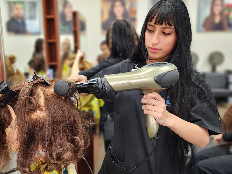 Cosmetology student in a black apron blow-drying a mannequin's hair in a salon classroom, with mirrors, styling stations, and other students in the background.