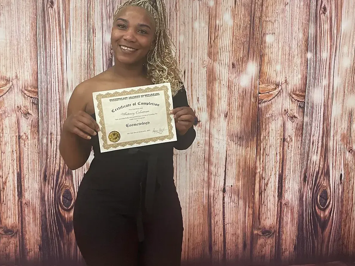 A smiling woman in a black outfit stands beneath balloons and a neon “CATA Academy” sign, holding a certificate in front of a wood-patterned backdrop.