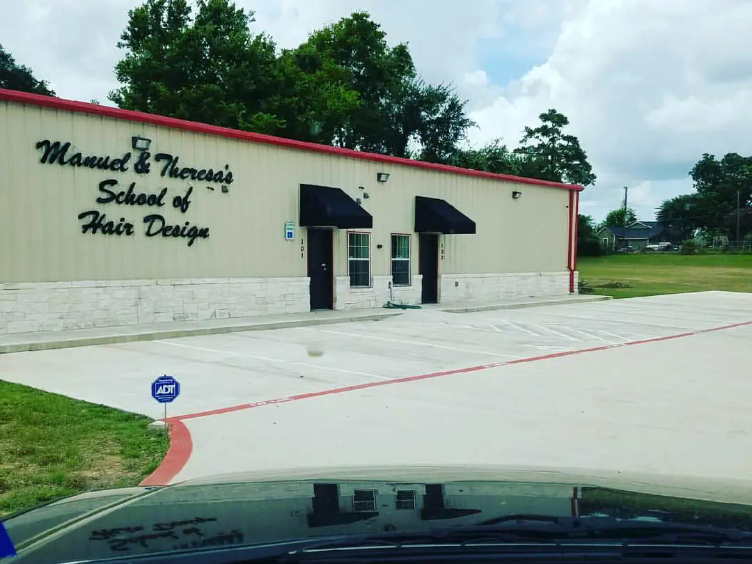 Single-story cream-colored Manuel & Theresa’s School of Hair Design building with red roof trim, two dark awnings, glass entry doors, a wide concrete parking area, and grass and trees beside it.