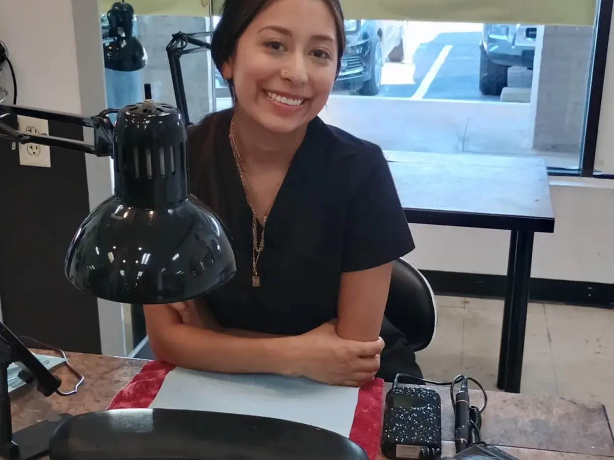 A smiling student in black sits at a manicure station with a black lamp, nail tools, and a red work surface beside large windows.