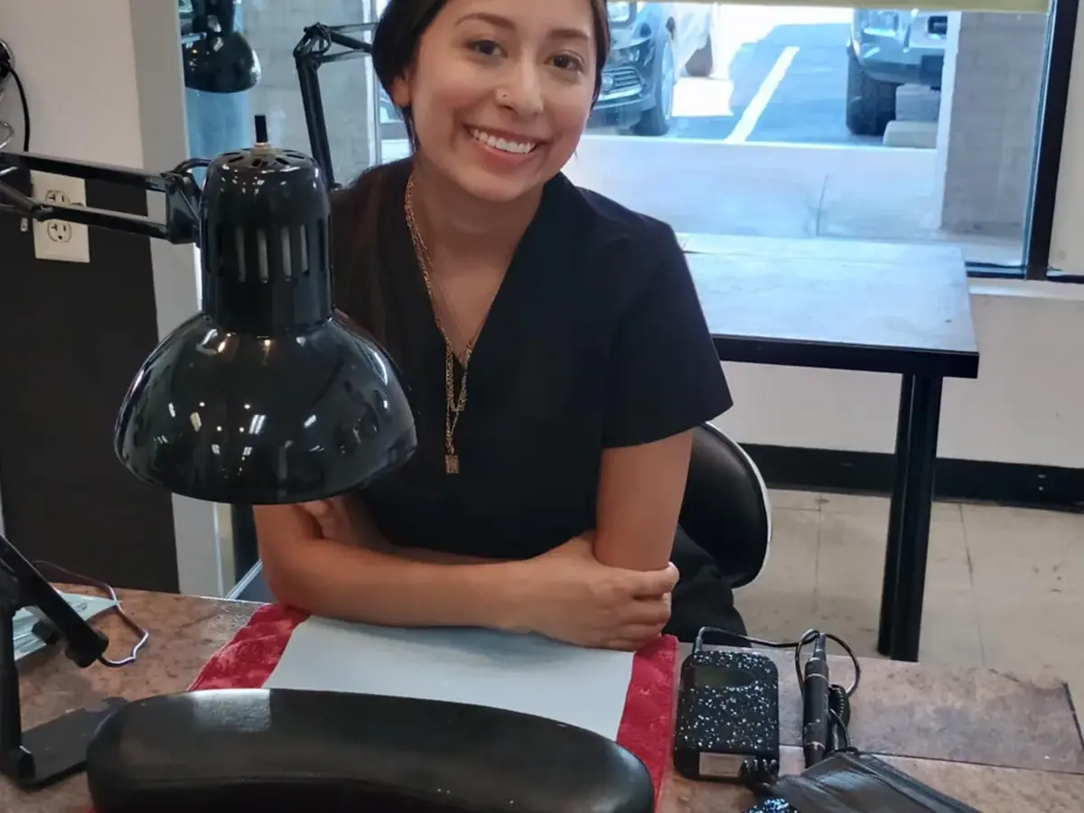Smiling student seated at a salon workstation beside a black lamp, with a window and parked cars behind her.