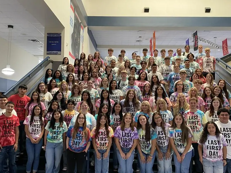 A large group of high school students in matching tie-dye shirts pose on the stairs and landing inside a school building, with flags and a balcony behind them.